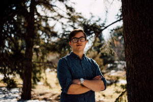 male student with glasses and blue button-up shirt stands underneath pine trees