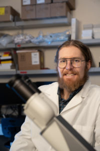 man with beard and glasses wearing a lab coat smiling in background behind microscope in foreground