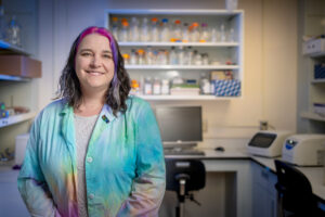 A woman with pink and purple colored streaks in her hair, wearing a rainbow tie-dyed lab coat, poses proudly for a photo in front of a section of a wet lab with countertop scientific equipment and open shelving full of beakers.