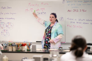 A woman with pink and purple colored streaks in her hair, wearing a rainbow tie-dyed lab coat, stands in front of a lab classroom facing her students. She is pointing up to the whiteboard behind her, which lists an mathematical equation for calculating molarity.