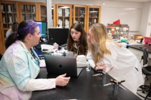 A woman with pink and purple colored streaks in her hair, wearing a rainbow tie-dyed lab coat, sits with her laptop facing two undergraduate students wearing lab coats. who are leaning in to discuss what is on the laptop screen in the wet lab classroom.