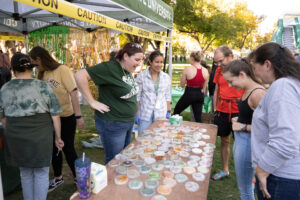Three individuals are standing in front of a table covered with colorful petri dishes full of agar art, as a woman in a green CSU shirt proudly talks about the display under a pop-up tent outside on the CSU Oval.