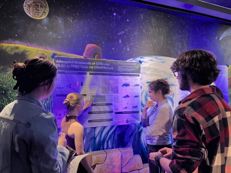 Students looking at presenter pointing at their poster, illuminated by a purple light