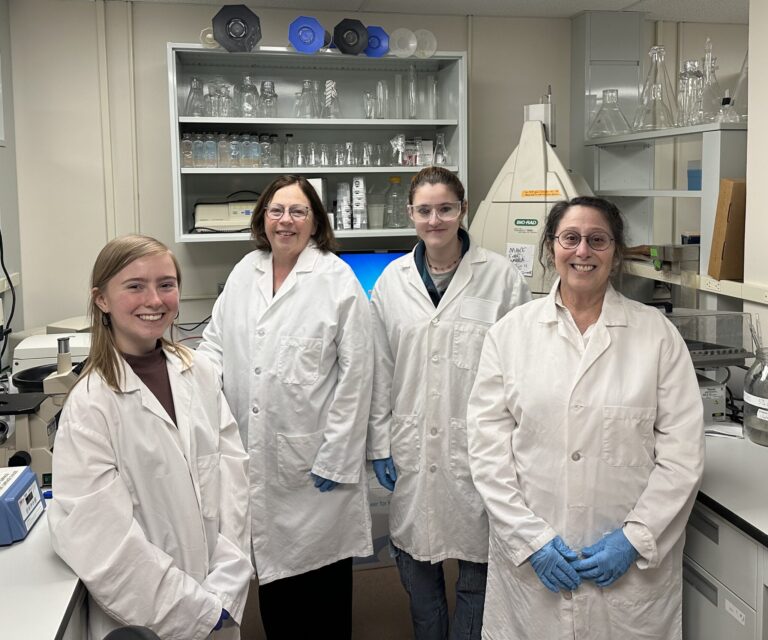 Four women in white lab coats pose in the lab